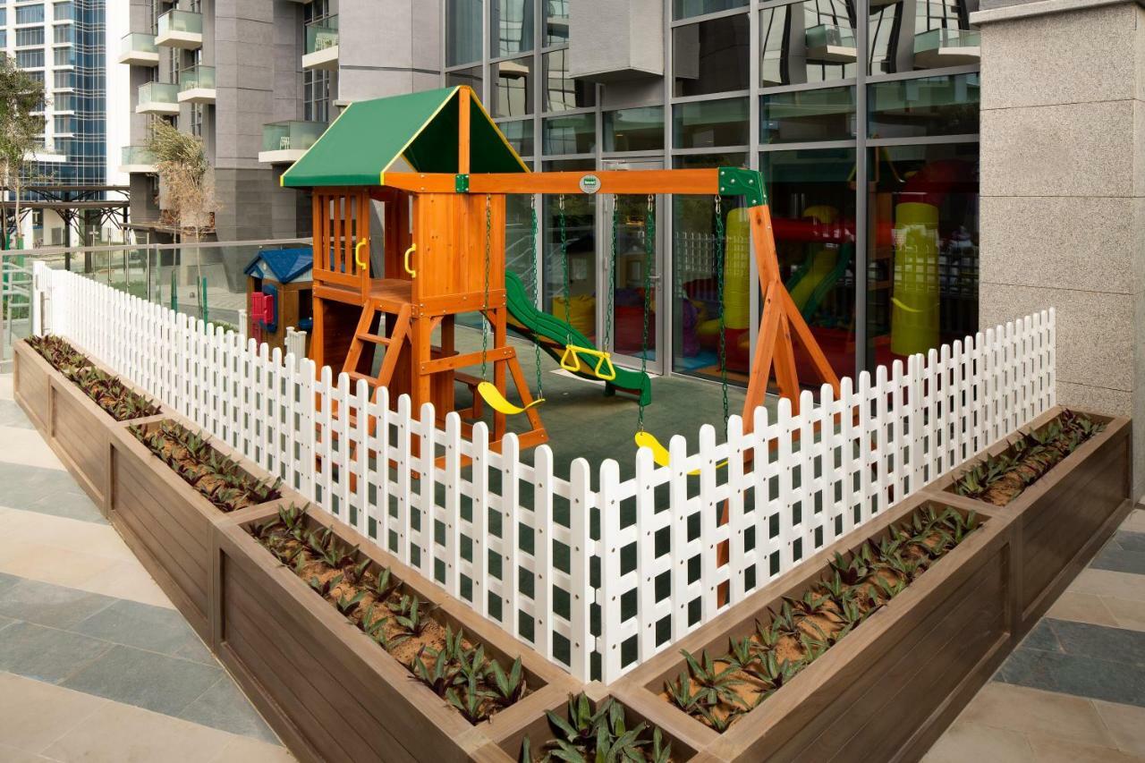 Millennium Atria Business Bay Aparthotel Dubai Ngoại thất bức ảnh The image shows a children's playground area, which is enclosed by a white picket fence. The playground features a wooden play structure with a slide, swings, and climbing elements. Surrounding the play area are planters with greenery, and the backdr