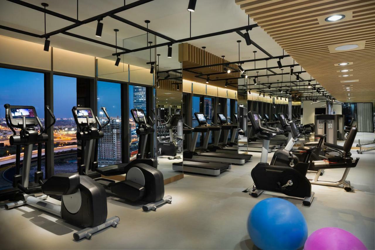 Millennium Atria Business Bay Aparthotel Dubai Ngoại thất bức ảnh The photo shows a modern gym with various types of exercise equipment available. There are several treadmills and elliptical machines arranged in rows, with large windows in the background providing a view of the city. The lighting is bright and cont