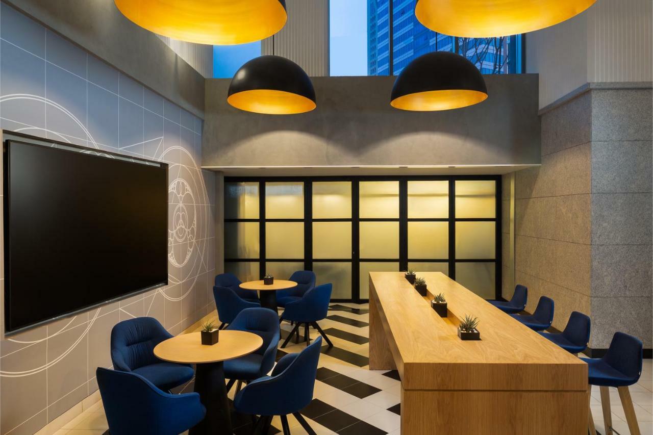 Millennium Atria Business Bay Aparthotel Dubai Ngoại thất bức ảnh The photo shows a modern interior space, likely a lounge or meeting area. It features a long wooden table with some small plants on it, accompanied by several blue, circular chairs arranged around it. There is a large flat-screen television mounted o