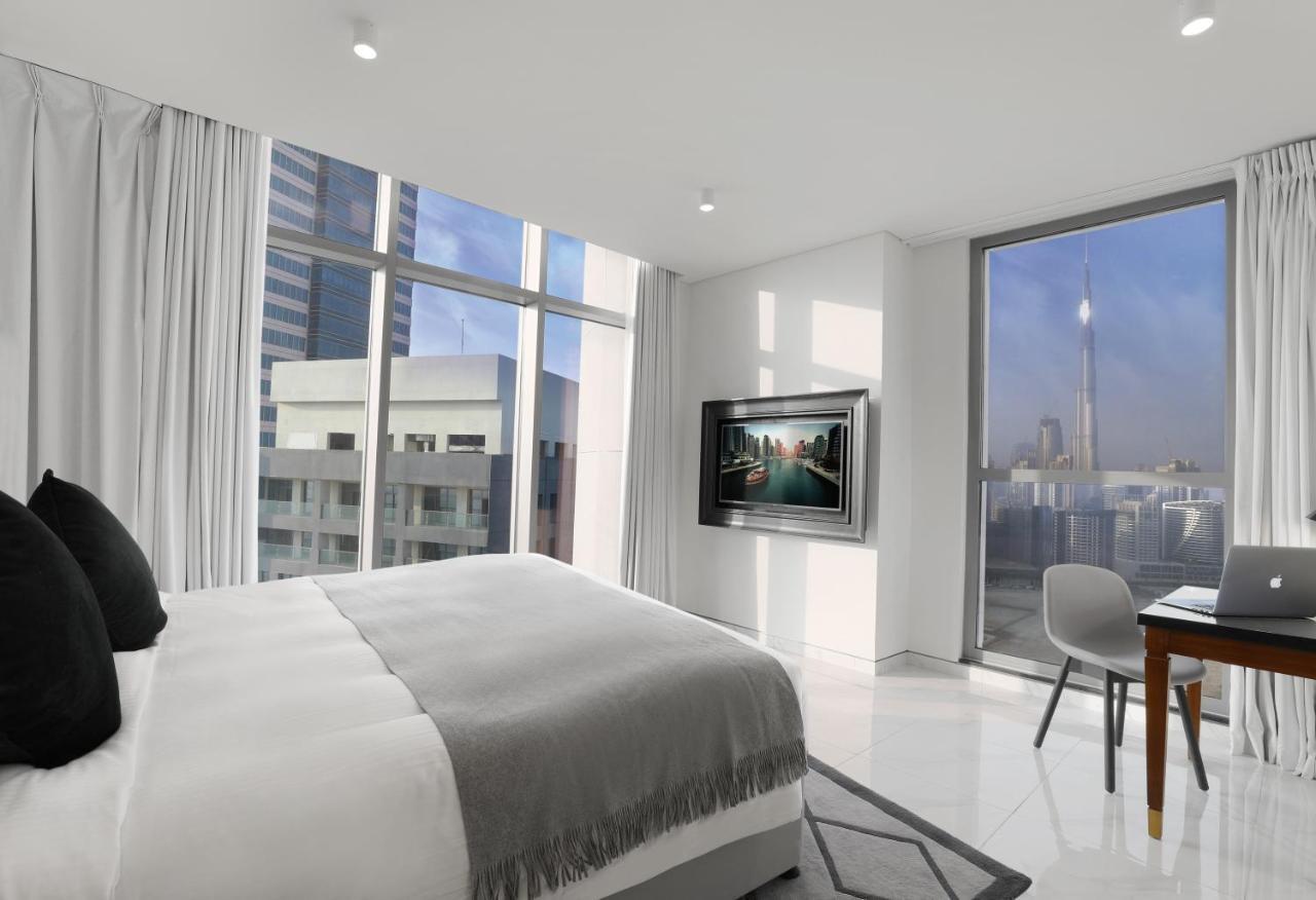 Millennium Atria Business Bay Aparthotel Dubai Ngoại thất bức ảnh The photo depicts a modern hotel room that features a large bed with a gray throw blanket. There are large windows allowing natural light to fill the space, showcasing a city skyline view. A flat-screen television is mounted on the wall opposite the 